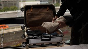 Man unpacking small suitcase, take out clothes and put on bed. Traveller check-in at hotel, move stuff out from bag. Plastic case lie on rack against window