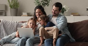 Smiling affectionate young family couple relaxing on weekend with small son and daughter, using computer software applications together, watching funny cartoons, web surfing or shopping online.