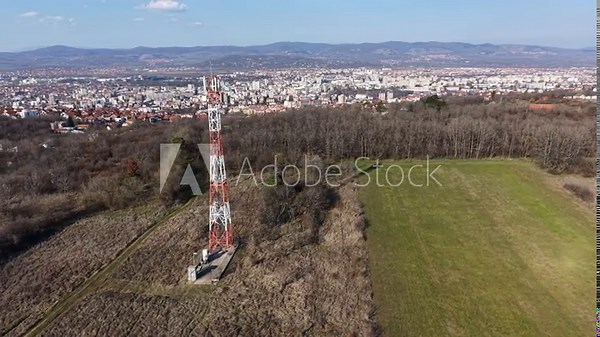 A view shows a telecommunication tower on a hill with open land and a city in the background