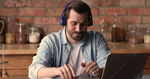 Young man sit in kitchen wear wireless headphones talk to foreign language tutor by video call, take part in English learning remotely. Modern tech, distant communication videoconference event concept