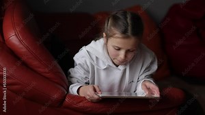 Curious cute teen girl using digital tablet technology device sitting on sofa. Little child holding pad computer surfing internet play game at home. Children tech addiction concept. Happy childhood.