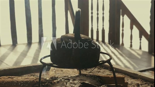 Weathered kettle placed on rustic stand over open fire against wooden veranda, showcasing warmth and simplicity of traditional tea brewing methods.