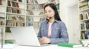 Asian female tutor in wireless headset giving video call training using laptop in university library. Young woman teacher coach has remote e-learning lesson for students, online course, talks remotely