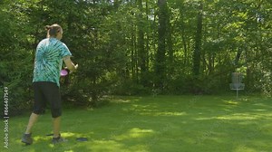 A man attempts a putt towards the basket in a wooded disc golf course, demonstrating concentration and skill in the sport