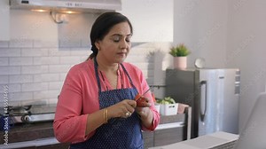 Indian Asian Matured Woman or Female wearing chef apron and standing in front of laptop in modern kitchen house setup chopping tomato while taking online cooking Tutorials courses to prepare recipe.