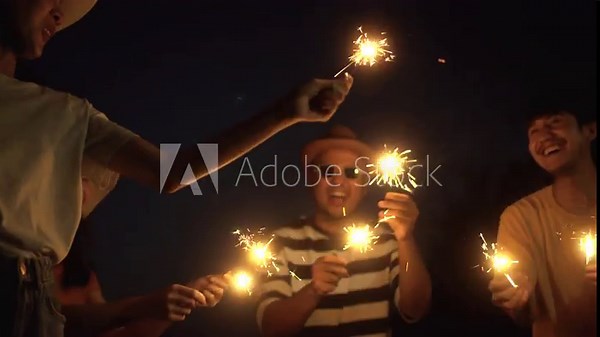 Happy friends group partying on the beach dancing and waving sparklers on summer at night. Group people having fun and enjoy on beach party smiling and dancing with sparklers.