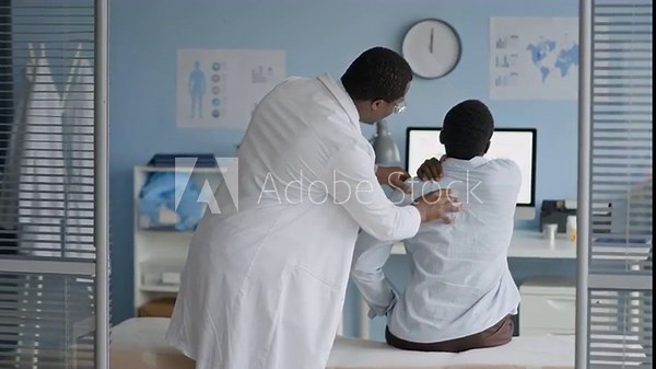 Medium shot of Black male physician performing physical examination on male patient upper back and shoulders, assessing posture, muscle tension, or spinal issues during clinical checkup