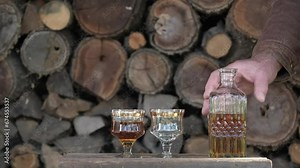 Cognac pouring in the glass from bottle. Hand puts a carafe with golden cognac and pouring into the glass, wooden table near logs. Strong drink - brandy or whiskey with ice. Alcohol amber drink Stock Video