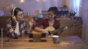 Group of female friends singing music while wearing headphones and playing guitar in warm modern cozy kitchen at home at night. happy girls love songs together happily with digital pad on wood table
