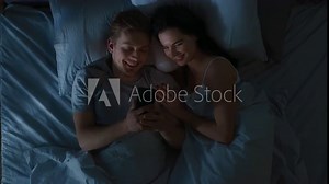 Happy Young Couple Use Smartphone in Bed at Night, They Share Screen, Browsing Through Social Networks, Sharing Pictures, Using Internet and Having Fun. Top View Camera Shot with Blue Colors