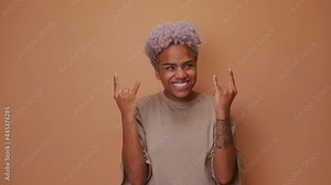 Positive joyful dark skinned woman makes heavy metal sign has fun on music rocking festival exclaims loudly closes eyes gestures actively, dressed in casual wear isolated over brown wall. Rock n roll.