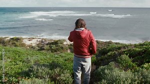 Rear view of couple of hikers venturing through a picturesque bushy landscape towards the stunning expanse of the ocean. Male and female travelers navigate their way down a scenic trail, surrounded by
