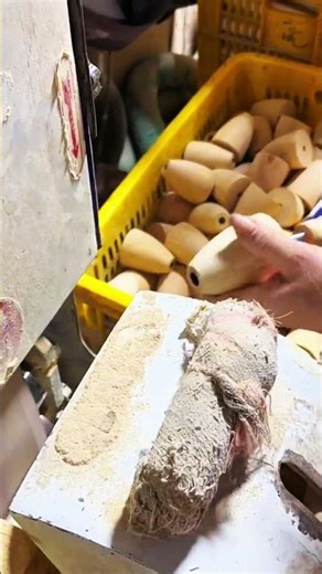 Smooth cylindrical wooden handle held near sawdust-covered machine, crate of similar pieces in