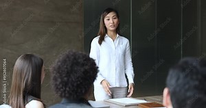 Serious young Asian business coach giving lecture, seminar, management class to diverse team. Project manager woman discussing creative ideas with colleagues, holding brainstorming meeting
