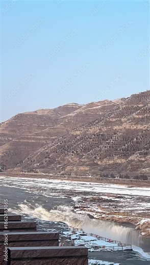 Linfen, Shanxi, China - January 2, 2026: A vertical video capturing the winter scenery of Hukou Waterfall. The Yellow River surges powerfully, creating a misty spray. The riverbanks are covered with s