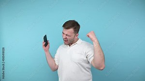 Angry man emotionally reacts to a loss with a smartphone in hand looks at the screen and screams in anger, isolated on a blue background