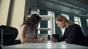 Two business women in office discuss topics, watching documents. blonde woman with straight hair, black shirt sits with paper, pen, speaking to employee, points on sheet of paper to colleague, looking