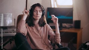 A beautiful girl with big headphones listens to music in a chair at the computer, dances, looks at the camera and smiles, the equalizer is playing on the monitor. Shakes his head to the rhythm