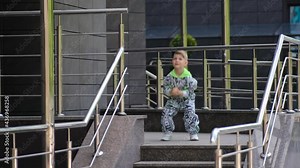 A small boy in a stylish suit dances a HIP-HOP dance near the entrance to the office building near the metal handrails. Modern dance in an urban street environment.