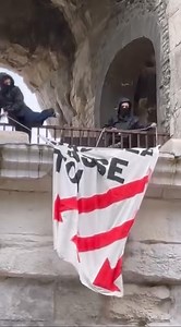 Nîmes. Deux femmes accrochent une banderole sur la façade des Arènes ce dimanche 2 après midi en face de l’établissement qui reçoit Jordan Bardella pour une séance de dédicaces de son livre. | Gazette Live Nîmes