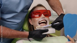Teeth shades, Dental procedure, Dental visit. Patient holds hand mirror as dentist, wearing mask and blue scrubs, compares tooth shades using shade guide.