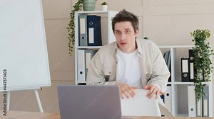 Funny caucasian man male millennial freelancer sitting down at desk in home office opens laptop screaming with fear scared frightened by image on screen hiding behind chair in horror closes computer