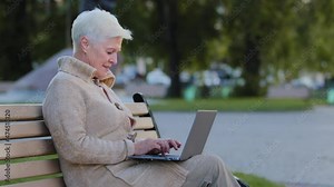 Senior gray-haired smiling woman of retirement age sitting on park bench using laptop. Granny retired typing on keyboard elderly lady browsing internet or working remotely communicating online with pc