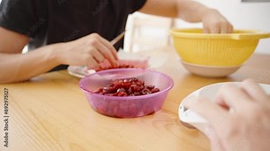 A Purple Bowl Filled with Pitted Cherries is being weighed. In the background, a person continues to pit cherries, focused on the task at hand. The yellow strainer and white bowl are ready for the nex