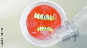 Concept shot above circular top view of glass filled with colourful drink, ice cubes & gold grills teeth sitting on top as liquid poured in from bottle creating fizz bubbles. Lean partydrink