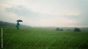 Woman in green raincoat fluttering in wind, walking through a misty green field with an open umbrella and bag,the wide landscape features scattered bushes, trees, and cloudy sky
