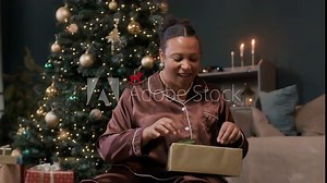 Young African American woman wearing pyjamas sitting in cozy living room opening Christmas gift
