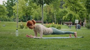 Concentrated caucasian woman standing in plank in the park on yoga mat. Outdoor plank exercising, training body strength during physical effort endurance. Morning sport routine.