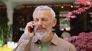 Happy european gray-haired older mid age bearded man talking on the phone outdoors. Smiling old senior adult male entrepreneur using cellphone making mobile call standing on city street outside.
