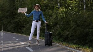 Joyful woman wait passing car in forest standing with suitcase and cardboard poster on roadside of highway. Blonde lady in hat jump from happiness to go anywhere. Travelling, hitchhiking, vacations