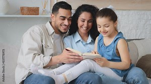 Hispanic multiethnic family sitting on couch sofa mother father and child girl reading book. Parents teach daughter to read literature novel story textbook help with homework home education knowledge