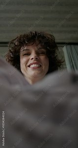 A woman in casual clothes opens a box and pulls out a shirt, view from the box. She's sitting on the couch at home. Daylight