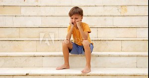 Young boy appears upset as he puffs his cheeks and rests his head on his fists. He raises his head slightly and gazes unhappily into camera, seemingly seeking words of encouragement or positivity.
