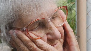 Portrait of grandmother with emotions and feelings. Old woman looking with sad expression outdoor. Grandma keeping hands at her face. Close up Side view Slow motion