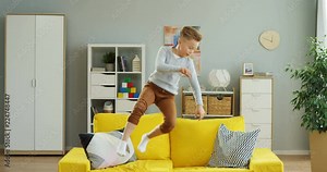 Cute little boy playing and jumping on the yellow couch in the living room, then falling on it and lying like sleeping because tired.