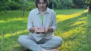 Charming young adult woman plays small musical instrument outdoor at sunrise. Balance concept. Female person relaxing meditates creating simple music sitting green landscape garden in golden sunlight Stock Video