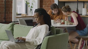Free stock video - Girl working on laptop computer sitting on sofa while her two roommates talking together sitting at kitchen table
