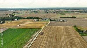 Aerial view of a patchwork of fields with a harvester in action. The fields are a mix of harvested and unharvested crops, with a road running through the landscape and farm buildings in the distance
