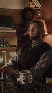 Vertical medium lowkey shot of young bearded Caucasian programmer working on software startup and showing progress on computer to his team, working and living together in 90s style garageVertical medi