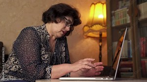 Senior woman in a glasses uses laptop, types text on a keyboard. She learns how to use computer. She is using social media. Laptop on the white table. Using technology in old age concept. Low angle