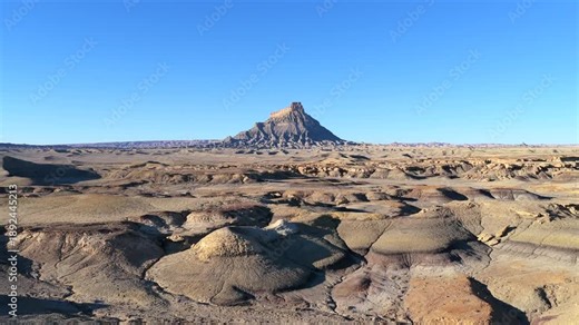 A wide landscape drone shot captures Factory Butte, a prominent geological feature in Utah. The isolated butte stands above a vast, barren desert and grey badlands terrain under a clear blue sky.