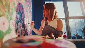 Professional female artist meticulously adding delicate details to vibrant floral painting, revealing creative passion while working in bright sunlit studio workspace