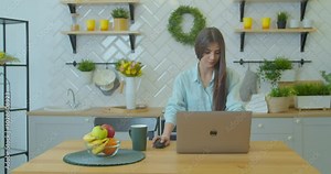 Attractive girl reading pleasant email, working remotely on computer in modern kitchen. Smiling young woman looking at laptop screen, answering messages, chatting with clients or friends.