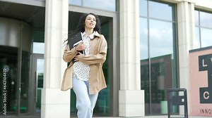Young female employee is running to a business meeting, going outside from the front door of an office building. Businesswoman with a folder is going to a conference. Brunette woman is late for work