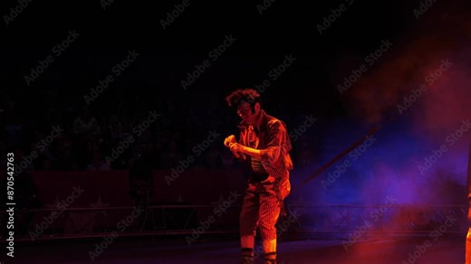 A clown with a funny hairstyle entertains guests in the circus arena in the light of colorful spotlights and smoke. He's tap dancing and singing into a microphone. Circus performance.
