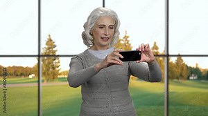 Smiling mature woman taking picture on her smartphone. Attractive senior woman taking selfie standing on windows background with beautiful nature outside.
